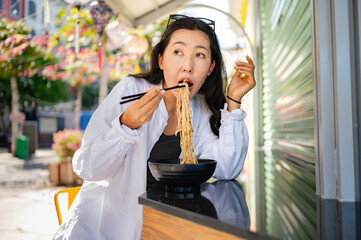 A young smiling Asian woman in a casual white shirt enjoys freshly prepared noodles with chopsticks at an outdoor café. She sits in a vibrant Asian street, focused and displaying a positive attitude