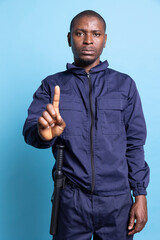 Serious security warden showing his authority with a restriction sign, limiting the access for people by raising a finger up in studio. Patrol agent in uniform does stop symbol as interdiction.