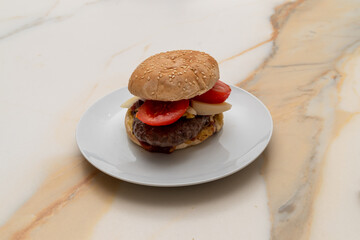 a homemade hamburger, with seeded bun, meat, tomato slices and cheese, with ketchup and mustard, on a white plate on a white marbled table