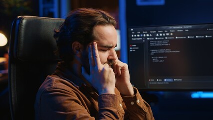 Annoyed software technician rubbing temples in frustration after failing to remove bugs appearing...