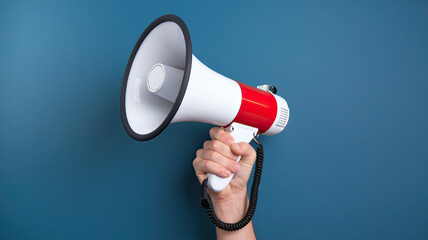 Hand Holding Red and White Megaphone Connected to Black Cord Against Blue Background, Communication Tool, Public Speaking, Announcement Device, Vibrant Colors, Clear Background