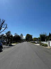 Fototapeta premium an empty residential street (driveways, houses, parked cars, trees, fences, gates) beneath a bright blue summer sky with clouds in Los Angeles - typical generic American suburb