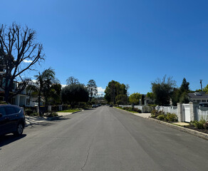 an empty residential street (driveways, houses, parked cars, trees, fences, gates) beneath a bright...