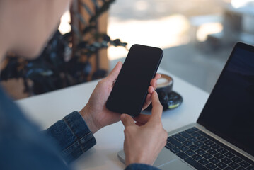 Cell phone black screen mockup, Woman hand holding mobile phone with blank screen while working at coffee shop. Business woman using smartphone, mockup for social media marketing