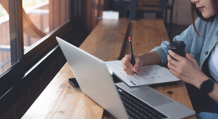 Business woman sitting on wooden table in coffee shop, drinking coffee, writing on notebook, working on laptop computer, close up. Female student studying online, e-learning, work from cafe