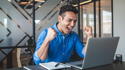 A man wearing and a blue-colored shirt, sitting at a table in what appears to be a modern office setting. He is excitedly looking at alaptop screen and has his right fist raised in a gesture of triump