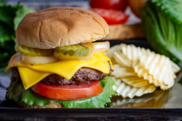 A cheeseburger on a black plate with pickles, fried onions, tomato an lettuce on a seedless bun.  Potato chips in the background.