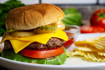 A cheeseburger on a white plate with pickles, fried onions, tomato an lettuce on a seedless bun.  Potato chips in the background.