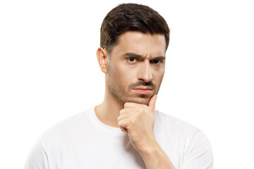 Fototapeta premium Close-up of young man in white t-shirt, holding his chin, looking at camera with expression of suspicion, mistrusting and doubting