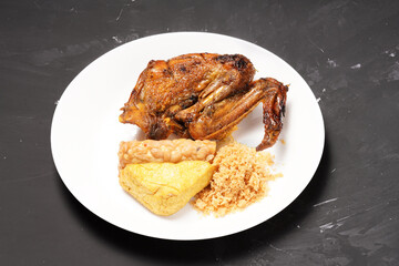 A plate of savory dishes including sweet grilled chicken with a dark brown color, fried tofu and tempeh, and crispy crumbs, served on a white plate against a dark background.