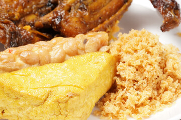 A plate of savory dishes including sweet grilled chicken with a dark brown color, fried tofu and tempeh, and crispy crumbs, served on a white plate against a dark background.