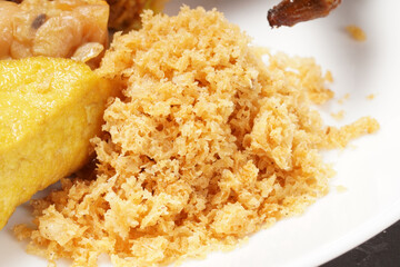 A plate of savory dishes including sweet grilled chicken with a dark brown color, fried tofu and tempeh, and crispy crumbs, served on a white plate against a dark background.