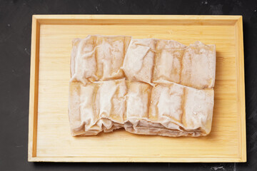 A vacuum-sealed plastic bag containing several frozen tofu bakso, square-shaped tofu with meatball filling tucked in the center, photographed on a wooden tray against a dark background.