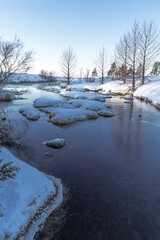 Wonderful morning winter view of a small Icelandic river with some trees and riverbanks covered with snow. Beautiful natural environment, peaceful background in Iceland. Snowy landscape with river.