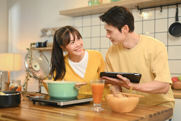 Happy young couple cooking together in a cozy kitchen and following a recipe on digital tablet