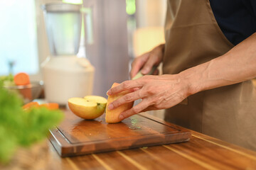 Man is cutting a fruit on a wooden cutting board, preparing healthy smoothie on countertop