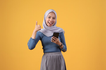 asian indonesian muslim woman holding smart phone giving thumb up finger gesture on isolated yellow background