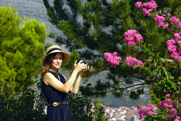 Smiling young woman in hat taking photo with DSLR camera standing in park on hill (Trebinje, Bosnia and Herzegovina). Portrait of female photographer during outdoor photo shoot on sunny summer day.