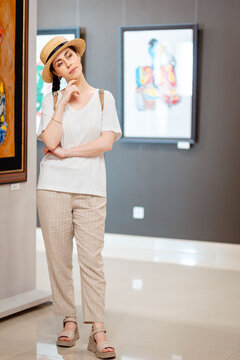 Historic education and museum day. Full height Pretty young Caucasian woman student wearing straw hat admiring paintings in the gallery. Vertical. Concept of cultural education