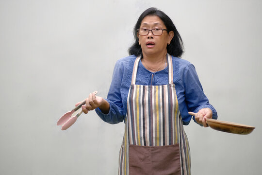 A woman wearing an apron holds a frying pan in one hand and food tongs in the other; confused expression