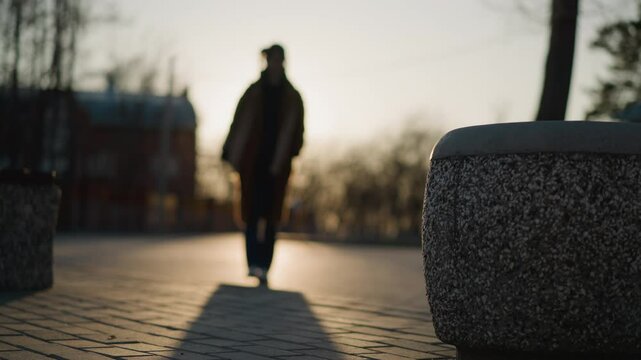 In a park at sunset, the silhouette of a person walking slowly in grief is visible, with the background and foreground both blurred, creating a soft, somber atmosphere