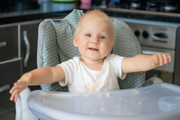 Cute blond caucasian baby child with blue eyes sitting at highchair after eating food. Child is dirty and everything is in rice grains. Baby is serious and surprised