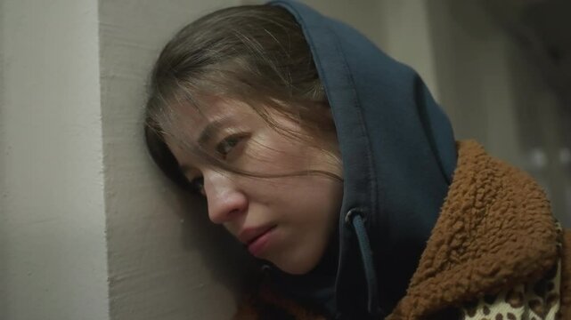 Close-up shot of a distressed girl in a brown coat, leaning against the wall of an underpass tunnel. She appears troubled, with a tense expression and her hoodie up, a mood of emotional turmoil