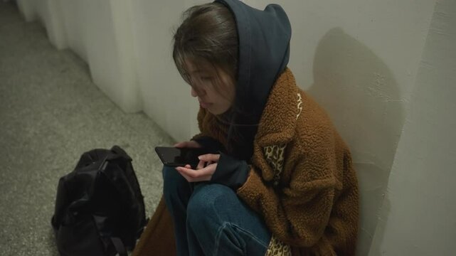 A girl in a brown coat sits bent over in an underpass tunnel, holding her phone with a distressed expression. She appears deeply troubled and lost in thought