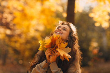 Smiling young woman enjoying the autumn season in the fall park with the yellow leaves at sunset