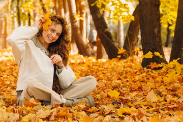 Smiling young woman sits in the autumn foliage on the ground of the forest with the yellow leaves