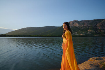 Woman in Vibrant Saree by a Scenic Mountain Lake, Serene Sunset with Woman in Traditional Attire by the Lake, Elegant Woman in Yellow Saree Posing by the Water's Edge Stock Photo.