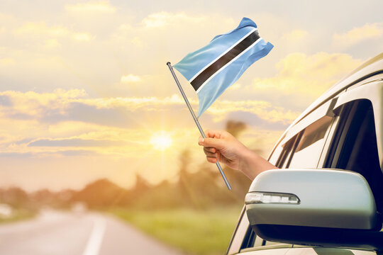 Waving the Botswana flag against the sunrise or sunset from a car driving along a country road. Holding the Botswana flag, traveling by car, on a weekend trip.