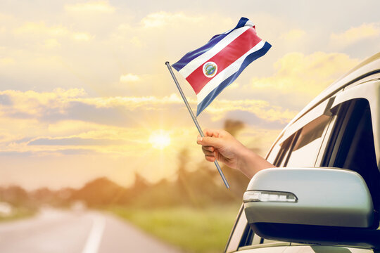Waving the Costa Rica flag against the sunrise or sunset from a car driving along a country road. Holding the Costa Rica flag, traveling by car, on a weekend trip.