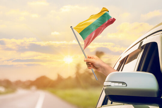 Waving the Lithuania flag against the sunrise or sunset from a car driving along a country road. Holding the Lithuania flag, traveling by car, on a weekend trip. - Powered by Adobe