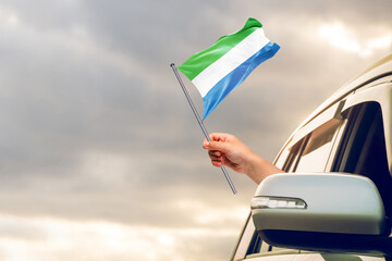 Waving the Sierra Leone flag against the sunrise or sunset from a car driving along a country road. Holding the Sierra Leone flag, traveling by car, on a weekend trip.