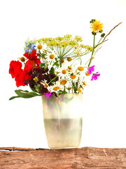 A vase filled with a colorful assortment of simple wild flowers sits on a wooden surface. The flowers include daisies, red flowers, and purple flowers. White background. Still life