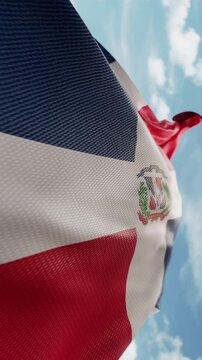 Wavy flag of Dominican Republic blowing in the wind in slow motion. Waving colorful flag team symbol abstract vertical background. Blue sky with clouds. World countries flying flags concept
