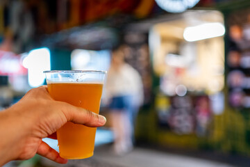 Close-up of a hand holding a glass of beer with a blurred background of a street market. Ideal for themes related to social gatherings, food and drink culture, or leisure activities.