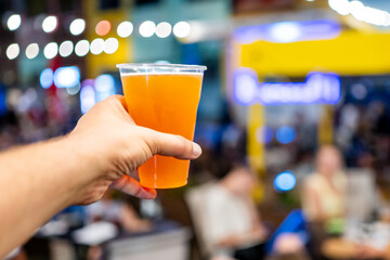A hand holding a glass of amber-colored beer with a blurred background featuring people and neon signs, creating a lively and vibrant atmosphere. Perfect for themes of nightlife, social gatherings