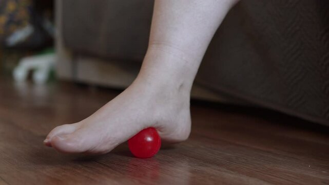 Unrecognizable woman rolls ball on floor with foot while sitting on sofa at home close-up. Person stretches leg, massages and treats flat feet. Prevention of hallux valgus.