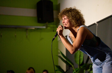 Caucasian female performer with curly hair delivering a monologue on stage in a dimly lit theater with audience