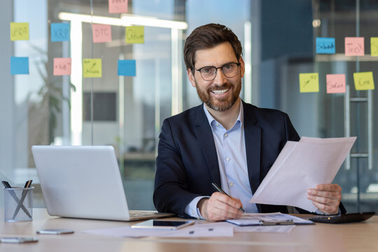 Confident businessman with glasses seated at desk reviewing documents. Smiling while holding paper, surrounded by modern office environment. Notes on glass wall suggest planning and organization.