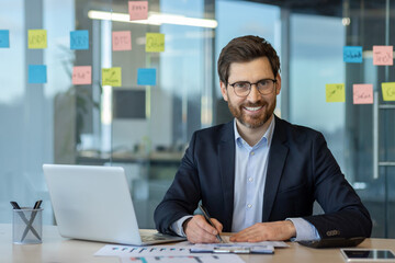 Confident businessman sitting at desk in modern office, using laptop and writing notes....