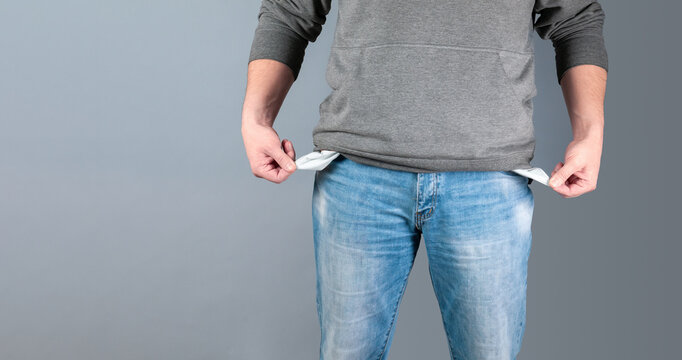 Young man in blue jeans and hoodie showing empty pockets on grey studio background. Millennial guy losing money, facing bankruptcy or unemployment during financial crisis. Economic depression concept