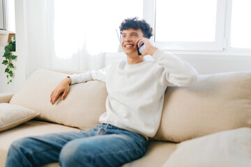 Fototapeta premium Smiling young man sitting on a couch talking on the phone, dressed in a white sweater and casual jeans, natural light illuminating the cozy living room environment
