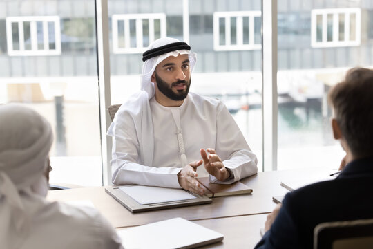 Serious young handsome Arab entrepreneur in traditional clothes talking to diverse partners at meeting, sitting at large table, speaking, discussing teamwork, cooperation, contract terms