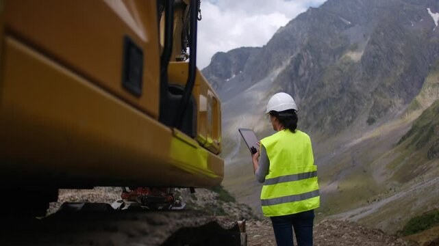 Female engineer with helmet using tablet near excavator at construction sit