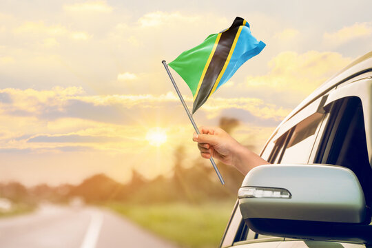 Waving the Tanzania flag against the sunrise or sunset from a car driving along a country road. Holding the Tanzania flag, traveling by car, on a weekend trip.