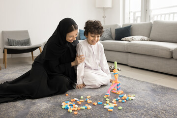 Happy Arabian child boy looking at toy tower on floor built with help and support of loving, caring...