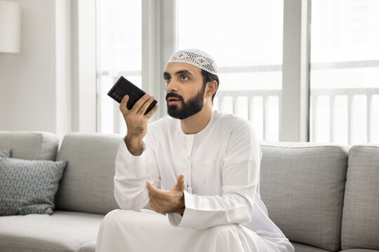 Serious bearded Muslim man in Arabic clothes talking on audio call on mobile phone, holding smartphone at mouth, speaking, using gadget for online communication at home - Powered by Adobe